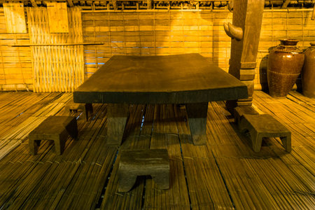 Hanoi, Vietnam - September 15, 2022: A large wooden table with four wooden stools is displayed on a bamboo floor inside a traditional Ede stilt house at the Museum of Ethnology, with bamboo walls and large ceramic jars visible in the background.のeditorial素材