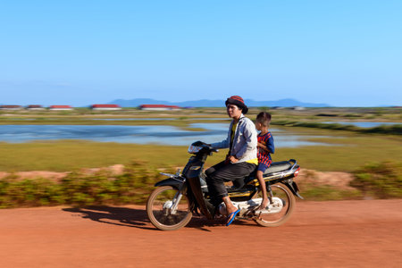Marais salants de Kampot, Cambodia - January 27, 2020: A man and a child ride together on a motorcycle along a dirt road, with salt marshes, water, and distant buildings visible in the background under a clear blue sky.のeditorial素材