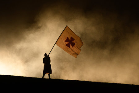 Unknown location - July 17, 2021: A person in costume is silhouetted against stage smoke while holding a large flag with a cross symbol during a nighttime outdoor performance.のeditorial素材