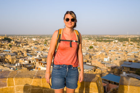 Jaisalmer, India - March 27, 2022: A woman wearing sunglasses, a red sleeveless shirt, denim shorts, and a backpack stands on a stone wall with a panoramic view of densely packed yellow sandstone buildings and rooftops in Jaisalmer under clear daylight.のeditorial素材