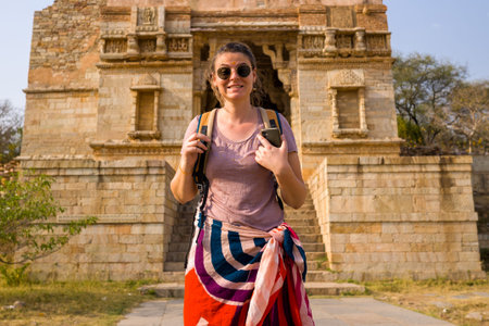 Chittorgarh, India - March 21, 2022: A person wearing sunglasses and a backpack stands on a stone path in front of the ruins of an ancient stone gateway with steps leading up to the entrance at Place Johar, with weathered walls and architectural details vのeditorial素材
