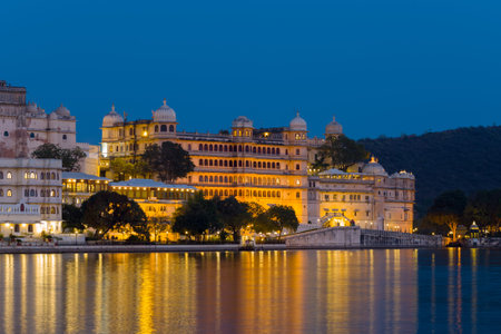 Udaipur, India - March 21, 2022: The image shows City Palace brightly illuminated at night, with its domed pavilions and ornate facade reflected in the calm waters of Lake Pichola under a deep blue sky.のeditorial素材