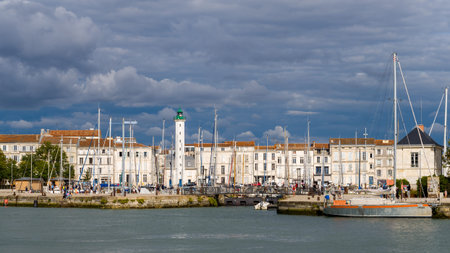 La Rochelle, France - August 30, 2020: A white lighthouse with a green top stands among moored sailboats along the harbor, with historic stone buildings and numerous people visible on the quay under a cloudy sky.のeditorial素材