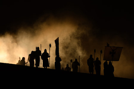 Unknown location - July 17, 2021: A group of people in costume are silhouetted against stage smoke while holding medieval-style flags, including one with a cross symbol, and polearms during a nighttime outdoor performance.のeditorial素材