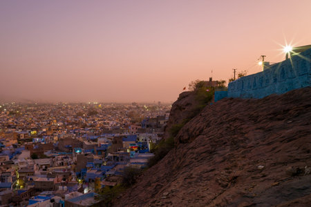 Jodhpur, India - March 26, 2022: The densely packed blue-painted houses of Jodhpur are visible below a rocky hillside at sunset, with a blue-painted wall and city lights beginning to appear in the distance.のeditorial素材