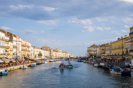 Sete, France - July 10, 2021: Crowds of people are seen gathered along both banks of the Canal Royal, with boats on the water and historic multi-story buildings lining the canal under a partly cloudy sky.のeditorial素材