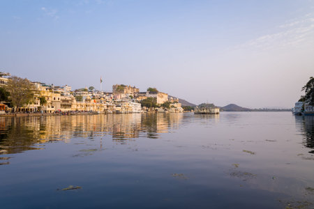 Udaipur, India - March 21, 2022: The image shows City Palace and a row of historic yellow and white buildings along the shore of Lake Pichola, with their reflections visible on the calm water under a clear sky.のeditorial素材