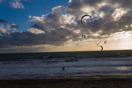 Le Havre, France - August 25, 2020: Multiple kitesurfers are riding waves on the sea with colorful kites in the air, under a dramatic cloudy sky at sunset, with a pebble beach in the foreground.のeditorial素材