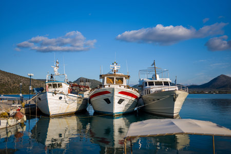 Port de Kilada, Greece - November 23, 2020: Three boats are moored together at the waterfront with calm water, hills, and partly cloudy sky in the background.のeditorial素材