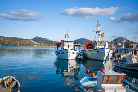 Port de Kilada, Greece - November 23, 2020: Several fishing boats are moored together at the waterfront with calm water, hills, and partly cloudy sky in the background.のeditorial素材