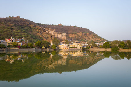 Bundi, India - March 20, 2022: Garh Palace is seen on a hillside overlooking Nawal Sagar Lake, with reflections of the palace, city buildings, and trees visible in the water under a clear sky.のeditorial素材