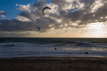 Le Havre, France - August 25, 2020: Several kitesurfers are visible on the sea with colorful kites in the air, riding waves near a pebble beach under a dramatic cloudy sky at sunset.のeditorial素材