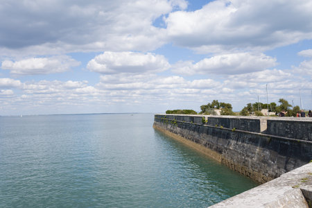 Ile de Re, France - September 1, 2020: A long stone seawall extends along the edge of calm coastal waters with a few people visible walking near the top of the wall under a partly cloudy sky.のeditorial素材