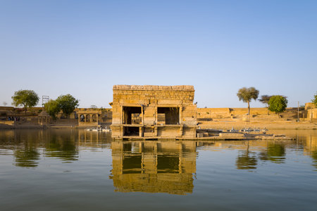 Jaisalmer, India - March 28, 2022: A rectangular sandstone structure stands partially surrounded by water at Gadisar Lake, with its reflection visible on the calm surface and trees, steps, and embankment along the lakeshore in the background.のeditorial素材