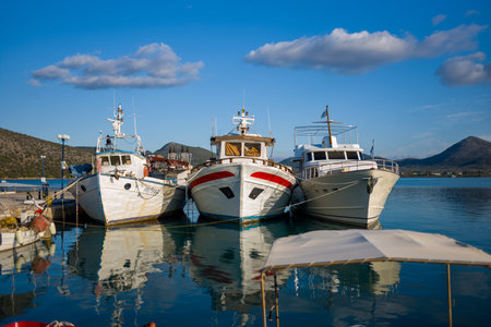 Port de Kilada, Greece - November 23, 2020: Three boats are moored together at the waterfront with calm water, hills, and partly cloudy sky in the background.のeditorial素材
