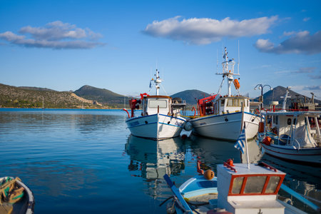 Port de Kilada, Greece - November 23, 2020: Several fishing boats are moored together at the waterfront with calm water, hills, and partly cloudy sky in the background.のeditorial素材