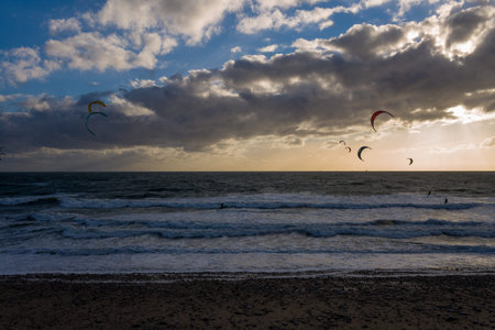 Le Havre, France - August 25, 2020: Several kitesurfers are visible riding waves on the sea under a cloudy sky at sunset, with colorful kites in the air and a pebble beach in the foreground.のeditorial素材