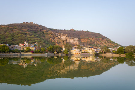 Bundi, India - March 20, 2022: Garh Palace is seen on a hillside overlooking Nawal Sagar Lake, with reflections of the palace, city buildings, and trees visible in the water under a clear sky.のeditorial素材