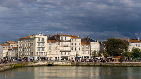 La Rochelle, France - August 30, 2020: Groups of people are gathered along the waterfront promenade with historic stone buildings, trees, and a bridge visible under a dramatic cloudy sky.のeditorial素材
