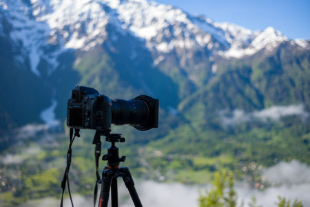 Unknown location - June 10, 2021: A digital camera with a telephoto lens is mounted on a tripod in the foreground, facing a green valley with scattered mist and snow-capped mountain peaks in the background, likely in the Alps.のeditorial素材