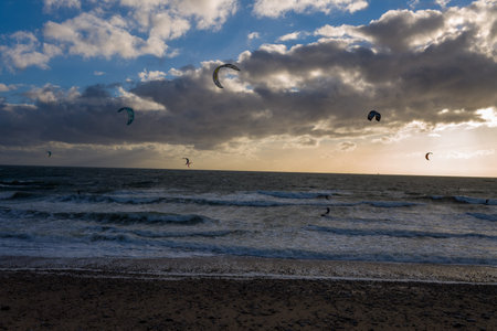 Le Havre, France - August 25, 2020: Several kitesurfers are visible on the sea with colorful kites in the air, riding waves near a pebble beach under a dramatic cloudy sky at sunset.のeditorial素材