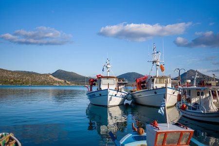 Port de Kilada, Greece - November 23, 2020: Several fishing boats are moored side by side at the waterfront with calm water, hills, and partly cloudy sky in the background.のeditorial素材