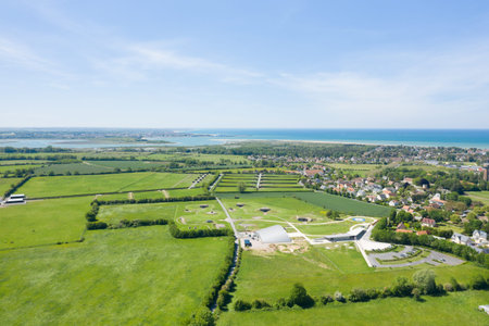Batteries de Merville, France - June 1, 2021: The image shows an aerial view of the Batteries de Merville, featuring preserved World War II bunkers, green fields, and a modern museum building with the town and coastline visible in the background.のeditorial素材