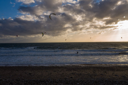 Le Havre, France - August 25, 2020: Multiple kitesurfers are riding waves on the sea with colorful kites in the air, under a dramatic cloudy sky at sunset, with a pebble beach in the foreground.のeditorial素材
