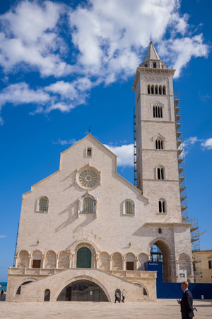 Trani, Italy - October 16, 2020: The facade of the historic stone Trani Cathedral with arched windows, a rose window, and an adjacent bell tower partially covered in scaffolding is visible under a blue sky, with a few people walking in the open square.のeditorial素材