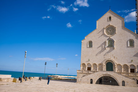 Trani, Italy - October 16, 2020: Several people are sitting and walking in the open square next to the large stone facade of a historic cathedral with arched windows and a rose window, located by the Adriatic Sea under a clear blue sky.のeditorial素材