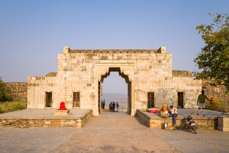 Chittorgarh, India - March 21, 2022: The Suraj Pole gate at Chittorgarh Fort is seen with several people nearby, religious statues and offerings placed on platforms, and a motorcycle parked at the entrance under a clear sky.のeditorial素材