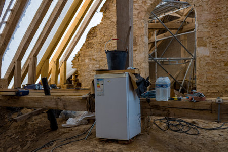 Eglise Saint Martin, France - July 26, 2020: A small refrigerator, black bucket, plastic water bottles, tools, exposed wooden beams, stone walls, and scaffolding are visible inside a roof renovation area at Eglise Saint Martin.のeditorial素材