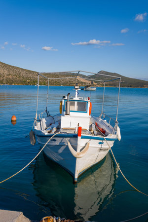 Port de Kilada, Greece - November 23, 2020: A white fishing boat is moored in clear blue water with hills and a partly cloudy sky in the background.のeditorial素材