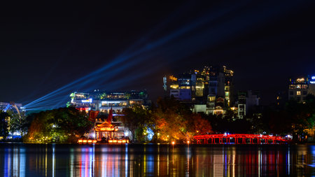 Hanoi, Vietnam - January 9, 2020: Ngoc Son Temple and The Huc Bridge are brightly illuminated at night on Hoan Kiem Lake, with colorful lights reflecting on the water and modern buildings with neon signage and spotlights visible in the background.のeditorial素材