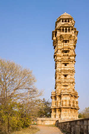 Chittorgarh, India - March 21, 2022: The image shows the full view of Vijay Stambh, also known as the Tower of Victory, a historic stone monument with intricate carvings, surrounded by dry trees and ancient stone structures under a clear sky.のeditorial素材