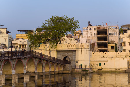 Udaipur, India - March 21, 2022: The image shows Daiji Bridge with its arched structure crossing the water, leading to a fortified yellow gateway and surrounded by historic multi-story buildings and a large tree, with reflections visible on the lake.のeditorial素材