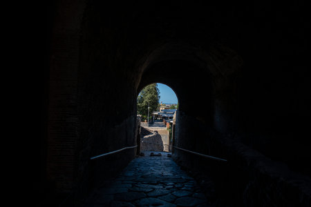 Pompei, Italy - October 4, 2020: The image shows a view from inside a dark ancient stone passageway looking out through an arched exit at Porta Marina, with sunlight illuminating the cobblestone path and ruins outside.のeditorial素材