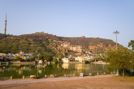 Bundi, India - March 20, 2022: Garh Palace is seen on a hillside overlooking Nawal Sagar Lake, with reflections of the palace, city buildings, and trees visible in the water under a clear sky.のeditorial素材