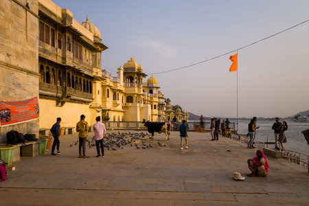 Udaipur, India - March 22, 2022: Several people stand and walk on the stone platform at Gangour Ghat by Lake Pichola, with pigeons, a cow, a seated woman in a pink sari, and historic buildings visible in the background under an orange flag.のeditorial素材