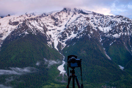 Unknown location - June 9, 2021: A digital camera mounted on a tripod is positioned in the foreground, facing towards a backdrop of snow-covered mountain peaks and forested slopes, likely in the Alps.のeditorial素材