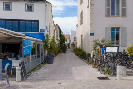 Ile de Re, France - September 1, 2020: A narrow pedestrian street lined with potted plants, bicycles, restaurant seating, and visible street signs is shown between white stone buildings under a partly cloudy sky.のeditorial素材