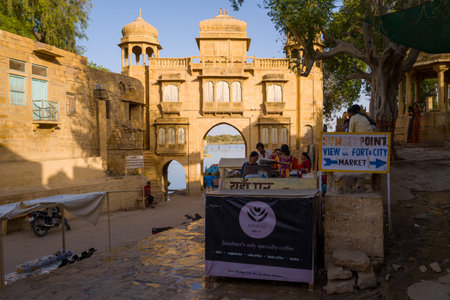 Jaisalmer, India - March 28, 2022: People are gathered at a coffee stall near the historic Tilon Ki Pol gateway leading to Gadisar Lake, with signage for a sunset point and market, motorcycles, and trees visible in the area.のeditorial素材