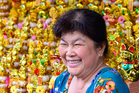 Ben Tre, Vietnam - January 17, 2020: A woman smiles in front of a market stall filled with numerous golden maneki-neko (lucky cat) figurines and other decorative items.のeditorial素材
