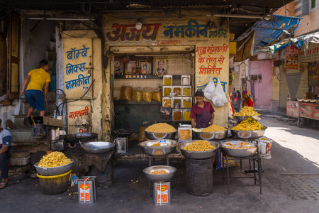 Udaipur, India - March 23, 2022: A street vendor stands behind large metal bowls filled with various snacks in front of a shop displaying Hindi signage, with additional people and commercial activity visible nearby during the daytime.のeditorial素材