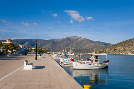 Port de Kilada, Greece - November 23, 2020: Fishing boats are moored along the quay beside a waterfront street lined with benches, palm trees, and buildings, with hills and wind turbines visible in the background under a clear blue sky.のeditorial素材