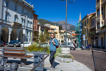 Ville de Omegna, Italy - September 28, 2020: An adult with sunglasses and a backpack stands near benches and a trash bin in a cobblestone square surrounded by multi-story buildings, parked cars, and mountains in the background under a clear blue sky.のeditorial素材