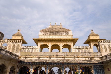 Udaipur, India - March 24, 2022: The ornate exterior of City Palace is shown with arched windows, decorative latticework, and domed pavilions under a partly cloudy sky, with people visible below the arches.のeditorial素材