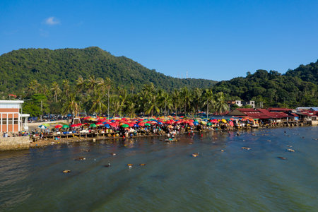 Marche de Kep, Cambodia - January 26, 2020: An aerial view shows a crowded waterfront seafood market with numerous colorful umbrellas, people, and market stalls along the shore, with palm trees and forested hills in the background.のeditorial素材