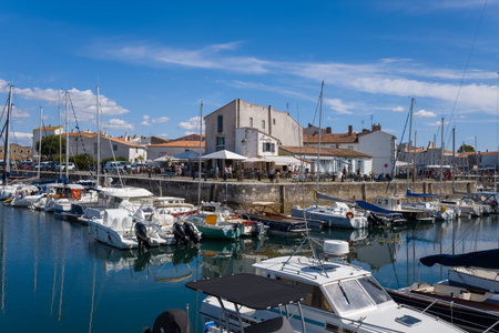 Ile de Re, France - September 1, 2020: Several sailboats and motorboats are moored in a calm harbor with white waterfront buildings, outdoor restaurants, and people visible along the stone quay under a blue sky with scattered clouds.のeditorial素材