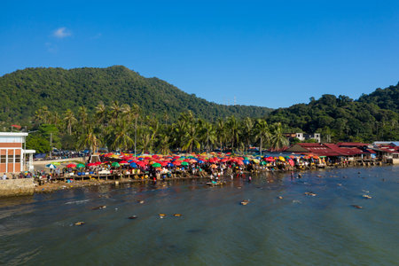 Marche de Kep, Cambodia - January 26, 2020: An aerial view shows a busy waterfront seafood market with numerous colorful umbrellas, people, and market stalls along the shore, with palm trees and forested hills in the background.のeditorial素材